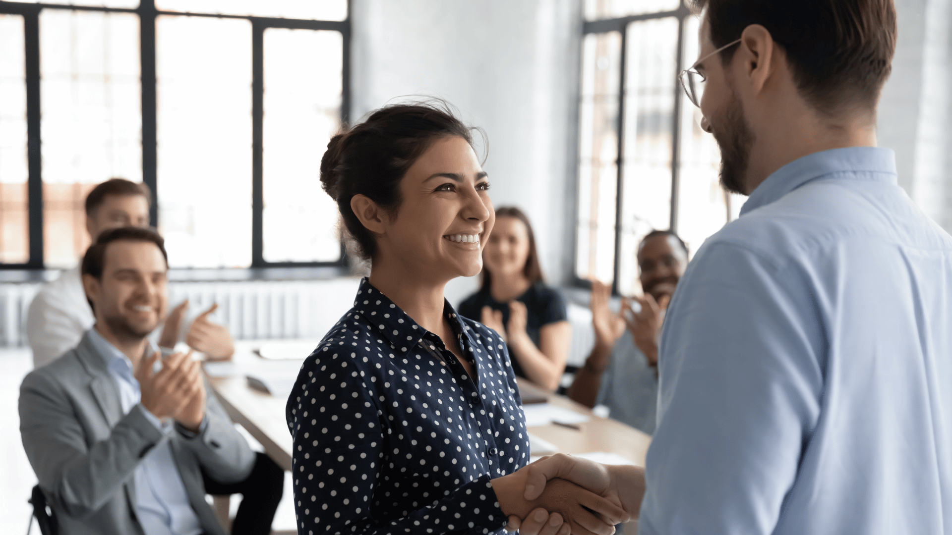 Employee getting promotion shaking hands with boss and receiving praises from colleagues in the background