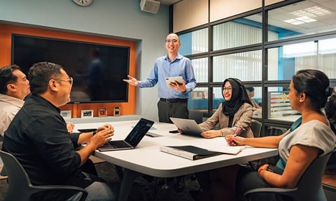 iPad Bald Man In Blue Shirt Holding Ipad Business Conference Room Presenting To A Group Of Colleagues Having A Discussion (720x432px)